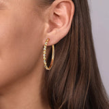 A close-up of a model's ear wearing a 14K gold hoop earring adorned with small clear diamonds, set against a soft gray background.