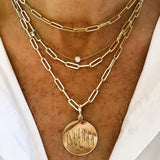 A close-up of a woman's neck wearing a layered gold necklace set. The set includes a paperclip chain, a delicate chain with a small round clear stone, and a round gold coin pendant engraved with calligraphy.