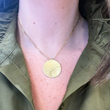 A close-up of a woman wearing a delicate gold chain necklace with a round, 14K gold gold pendant, styled with an olive green shirt.