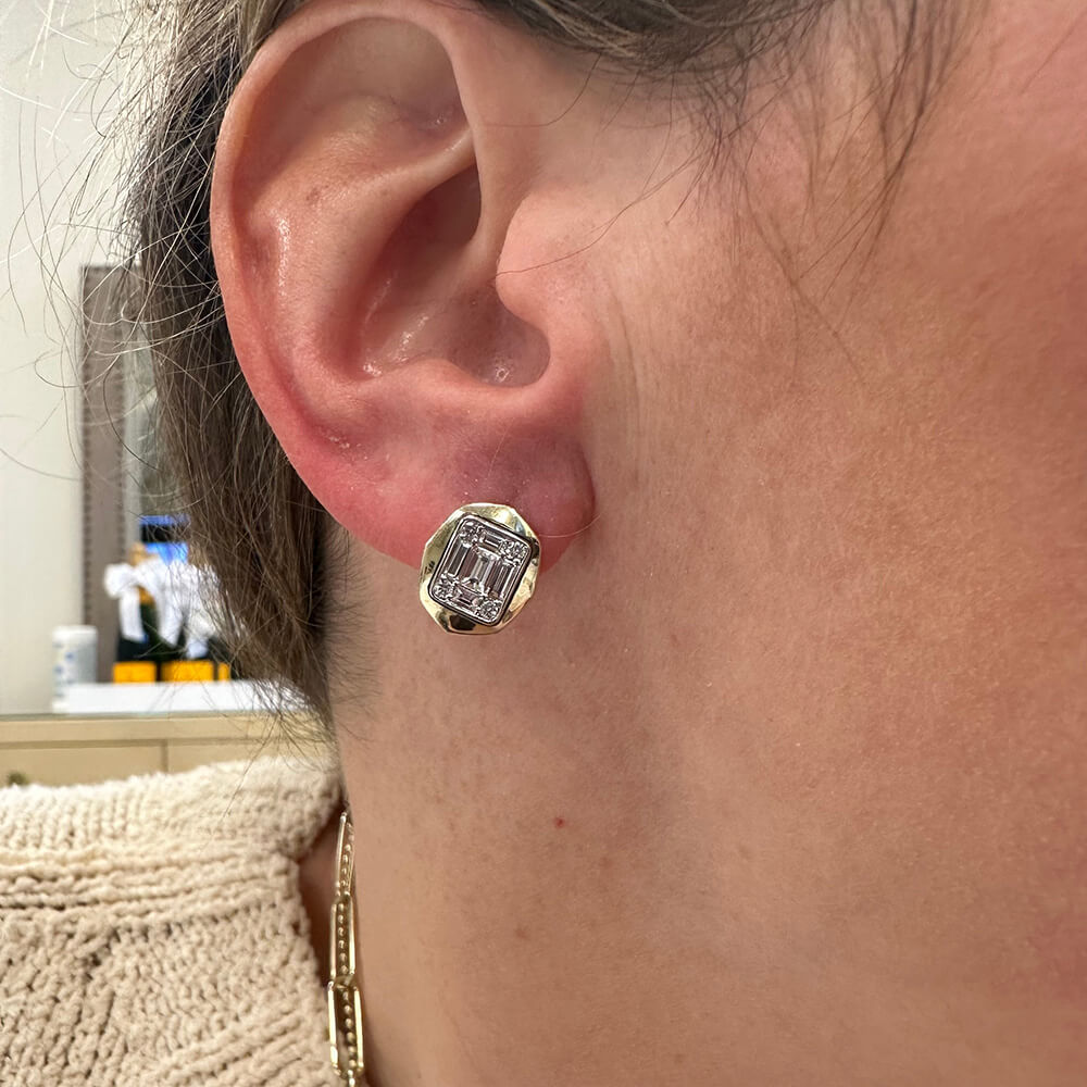A close-up of a woman's ear wearing a 14K gold stud earring with a clear rectangular diamond. ble behind them.