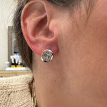 A close-up of a woman's ear wearing a 14K gold stud earring with a clear rectangular diamond. ble behind them.