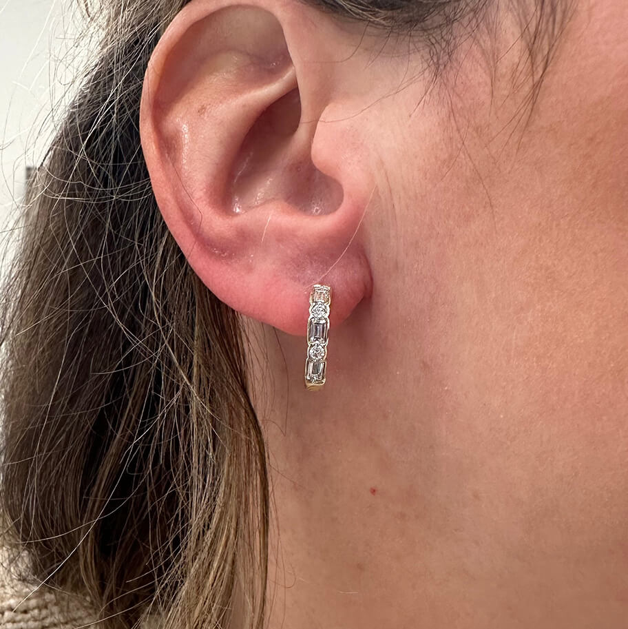 A close-up of a woman's ear wearing a 14K Gold huggie hoop earring with baguette-cut and round cut diamonds