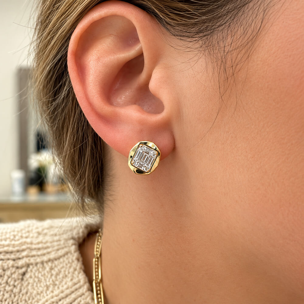 A close-up of a woman's ear wearing a 14K yellow gold stud earring with a clear rectangular baguette-cut diamond.