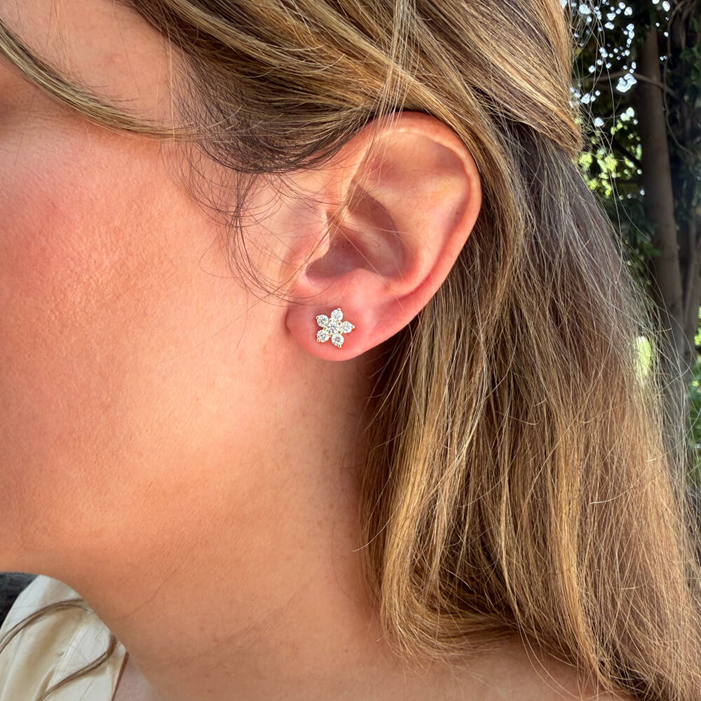 A close-up side view of a woman's ear wearing a 14K yellow gold diamond flower-shaped stud earring.