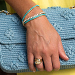 Hand wearing a gold ring and turquoise bracelet on a blue textured clutch against a yellow background