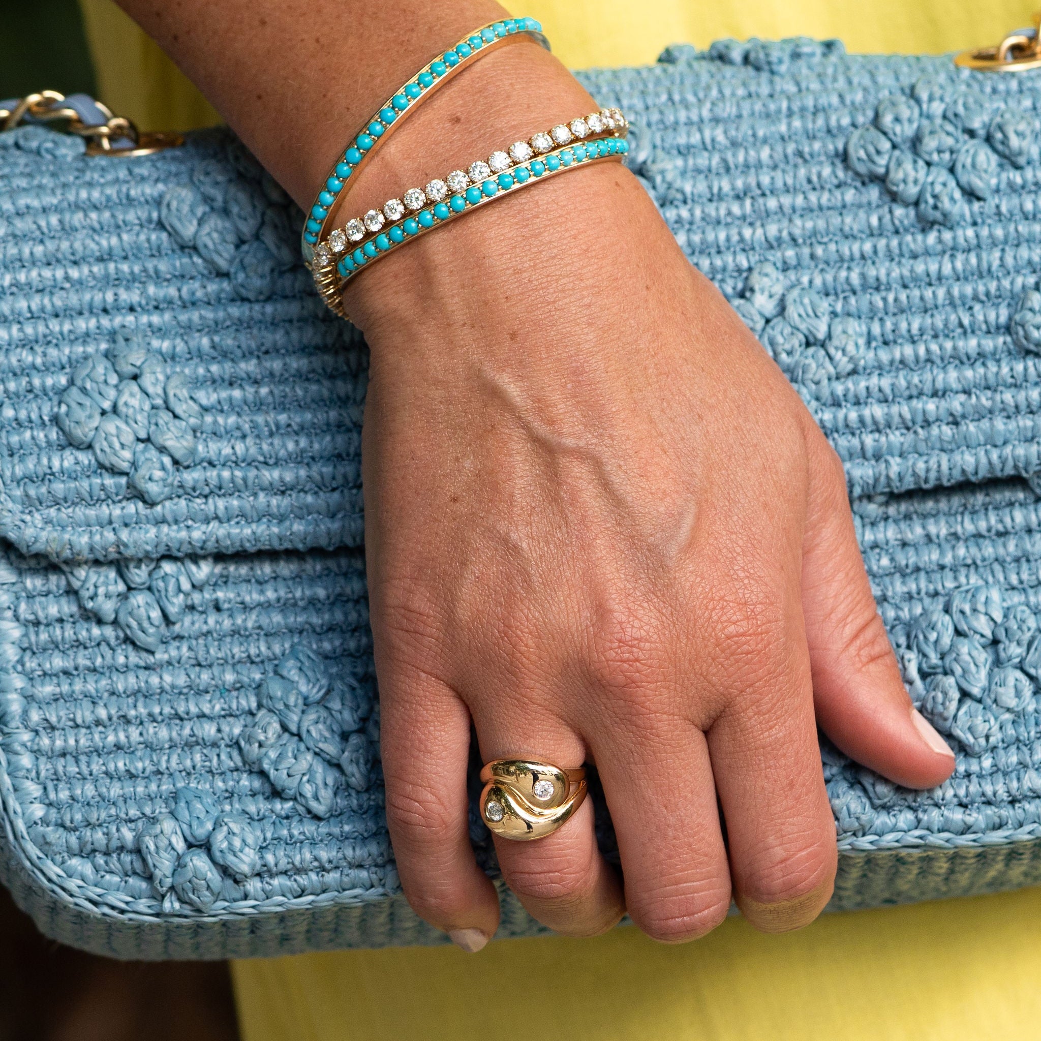 Hand wearing a gold ring and turquoise bracelet on a blue textured clutch against a yellow background