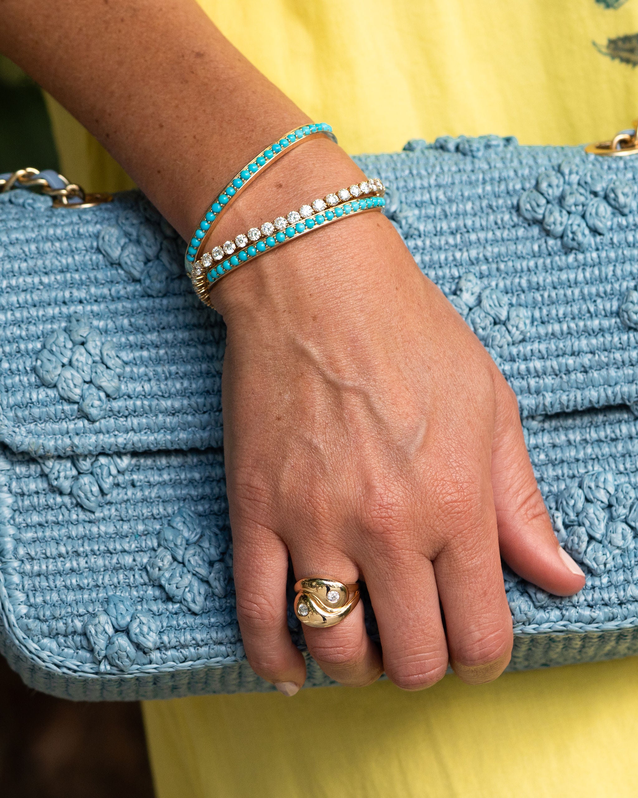 Hand wearing a gold ring and turquoise bracelet on a blue textured clutch against a yellow background