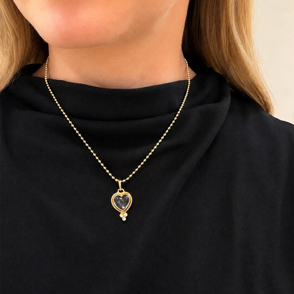 A close-up of a model wearing a delicate 14K yellow gold bead chain necklace with a heart-shaped pendant containing a dark, textured center and a small clear stone below it.