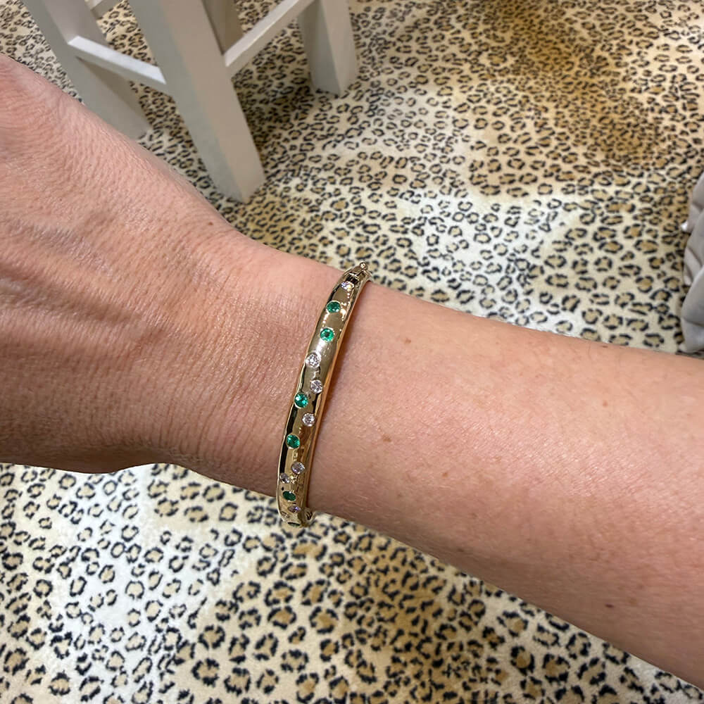 A close-up of a woman's wrist wearing a slim 14K gold bangle bracelet with emeralds and diamonds, set against a leopard-print rug 