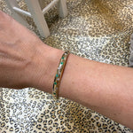 A close-up of a woman's wrist wearing a slim 14K gold bangle bracelet with emeralds and diamonds, set against a leopard-print rug 
