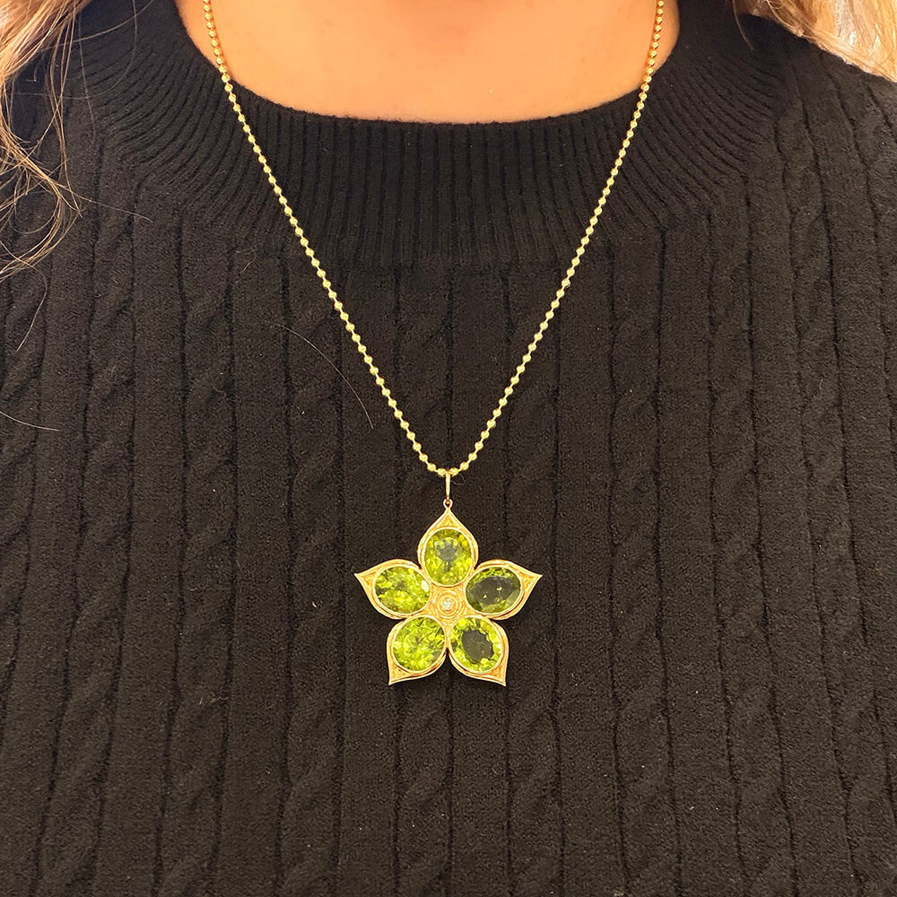 A 14K yellow gold flower-shaped pendant necklace with five green Peridot gemstone petals, worn by a person wearing a black cable-knit sweater.