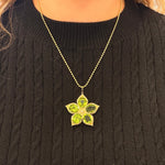 A 14K yellow gold flower-shaped pendant necklace with five green Peridot gemstone petals, worn by a person wearing a black cable-knit sweater.