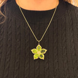 A 14K yellow gold flower-shaped pendant necklace with five green Peridot gemstone petals, worn by a person wearing a black cable-knit sweater.