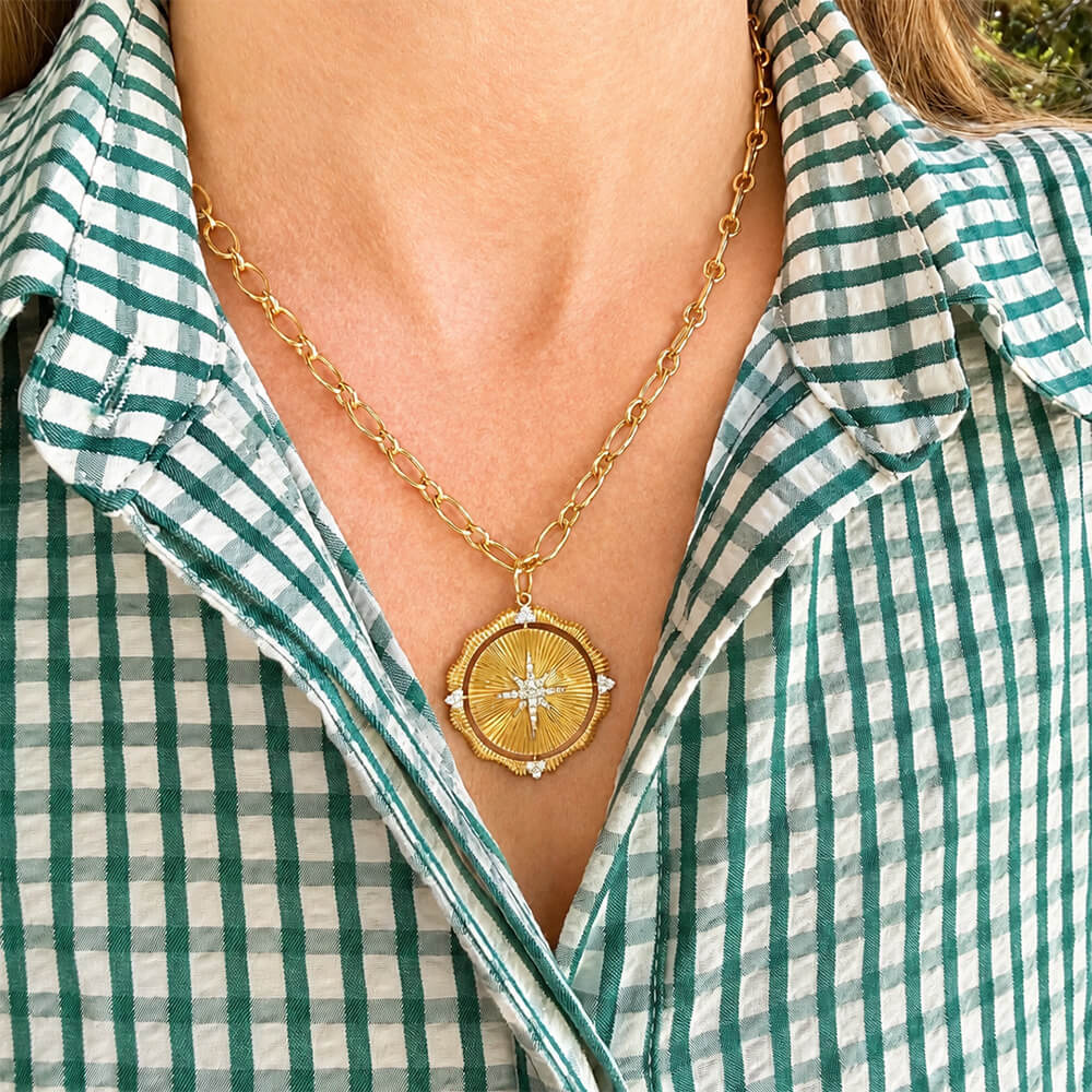 A gold necklace with a 14K gold circular compass pendant with diamonds is worn by a woman wearing a green and white gingham shirt.