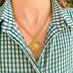 A gold necklace with a 14K gold circular compass pendant with diamonds is worn by a woman wearing a green and white gingham shirt.