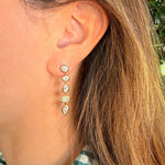 A close-up of a woman's ear wearing a gold drop earring with five clear faceted diamonds in a vertical arrangement.