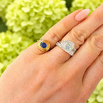 A close-up of a hand wearing two rings: an 18K yellow gold ring with a blue sapphire and a silver-toned diamond ring with an emerald-cut center stone, set against a soft-focus background of light green hydrangeas.