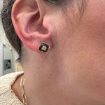 A close-up of a woman's ear wearing a small square stud earring with a single diamond surrounded by a gold beaded border.