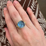 Hand wearing a gold ring with a blue topaz gemstone against a patterned fabric background