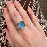 Hand wearing a gold ring with a blue topaz gemstone against a patterned fabric background
