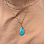 A 14K gold necklace with a teardrop-shaped turquoise pendant is worn around the neck over a dusty pink top.