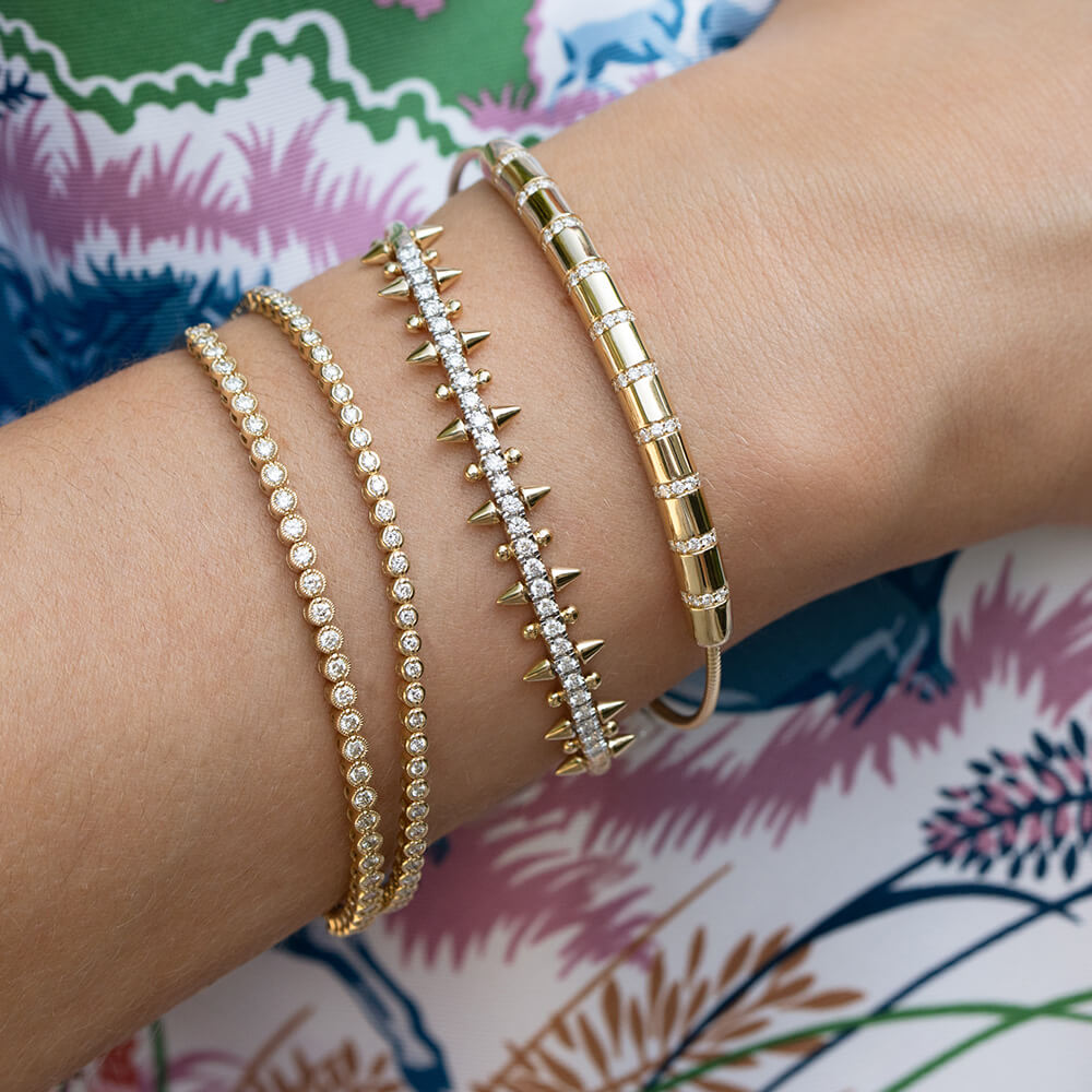 Four 14K yelllow gold bracelets with different designs (beaded, spiky, diamond-encrusted) stacked on a person's wrist against a colorful patterned background.