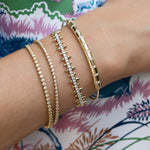 Four 14K yelllow gold bracelets with different designs (beaded, spiky, diamond-encrusted) stacked on a person's wrist against a colorful patterned background.