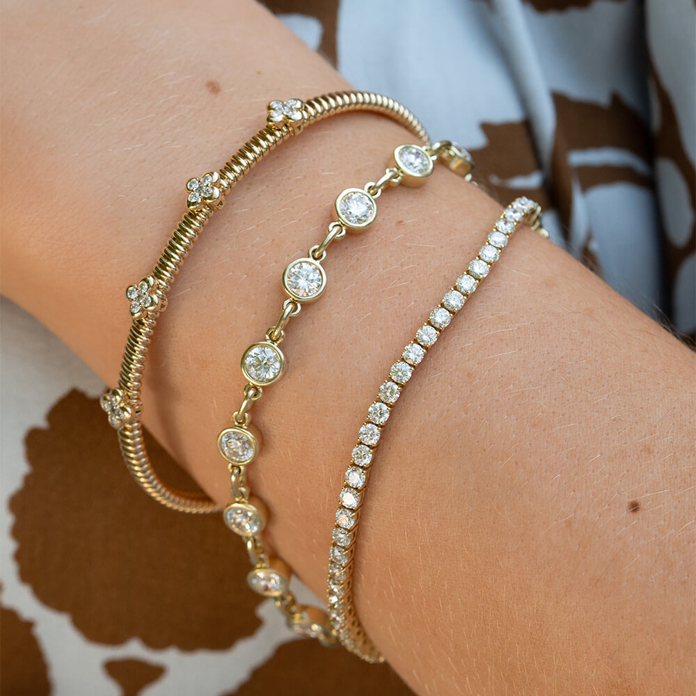 Three 14K yellow gold bracelets with clear diamonds worn stacked on a woman's wrist. The bracelets include a textured bangle, a chain with round diamonds, and a tennis-style link bracelet.
