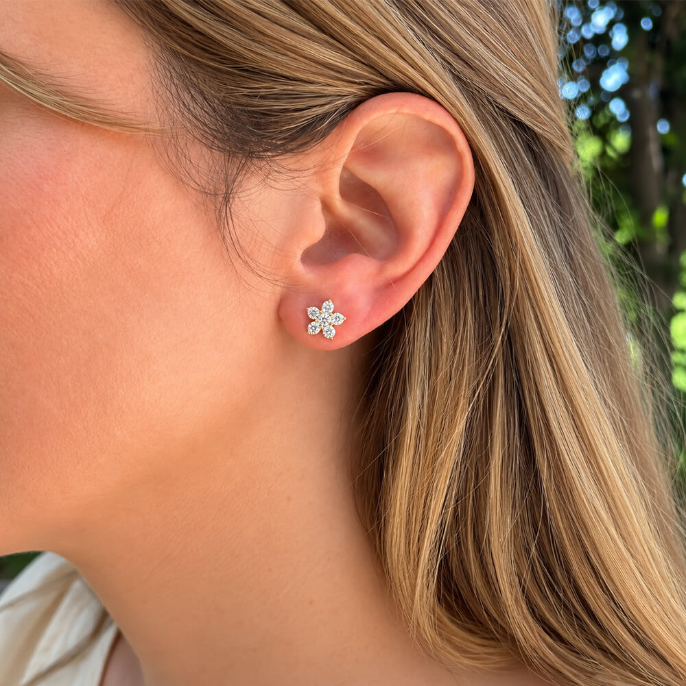 A close-up side profile of a woman wearing a 14k yellow gold flower stud earring with clear diamonds, set against her ear and blonde hair.