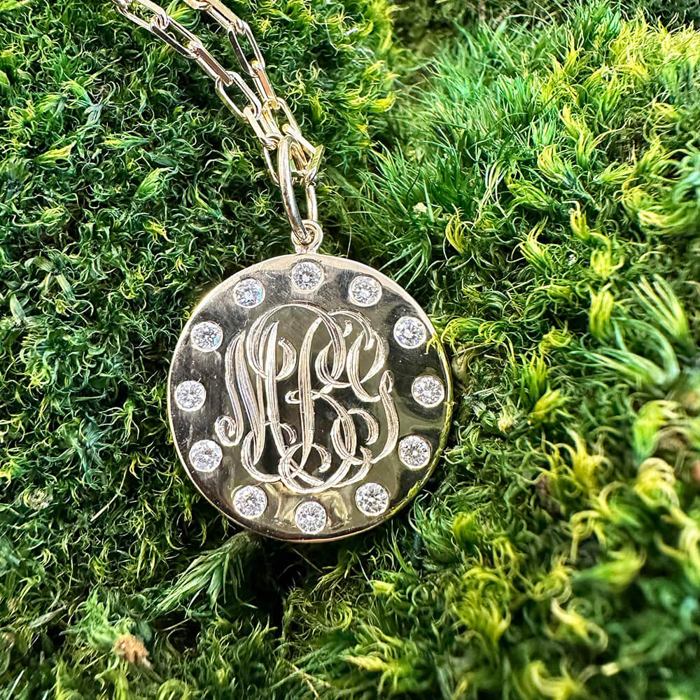A round gold pendant necklace with a monogram design and small clear diamonds, resting on a bed of green moss.