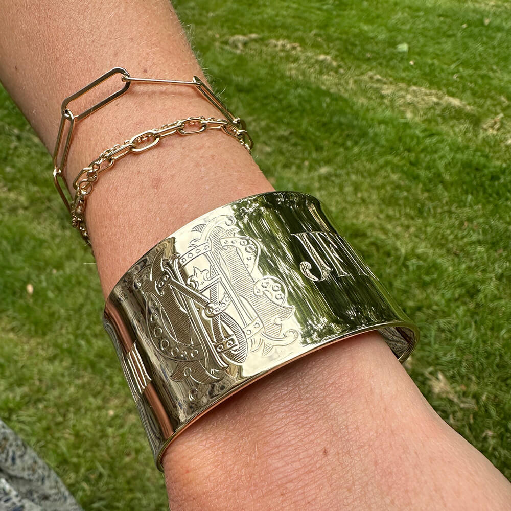 A close-up of a woman's wrist wearing a wide, polished gold cuff bracelet with engraved lettering and decorative motifs, along with two thinner gold chain bracelets, against a blurred green grass background.