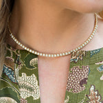 A close-up of a model wearing a 14K gold tennis necklace with small clear diamonds, paired with a green patterned blouse.
