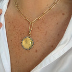 A gold chain necklace with a large 14K yellow gold circular pendant, worn on a woman's neck. The pendant features a polished gold center surrounded by a border of small dark blue sapphires, set against a white collared shirt.