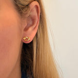A close-up of a woman's ear wearing a small 14K gold love knot earring against a plain light background.