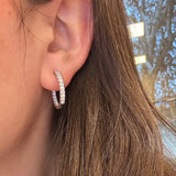 A close-up of a woman's ear wearing a 14K white gold hoop earring set with clear diamonds, with brown hair visible against a softly blurred outdoor background.