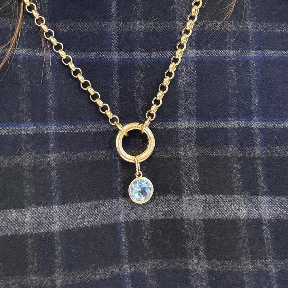 A gold chain necklace with a small gold bail and a dangling light blue topaz round pendant, displayed on a dark blue plaid fabric background.