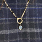 A gold chain necklace with a small gold bail and a dangling light blue topaz round pendant, displayed on a dark blue plaid fabric background.