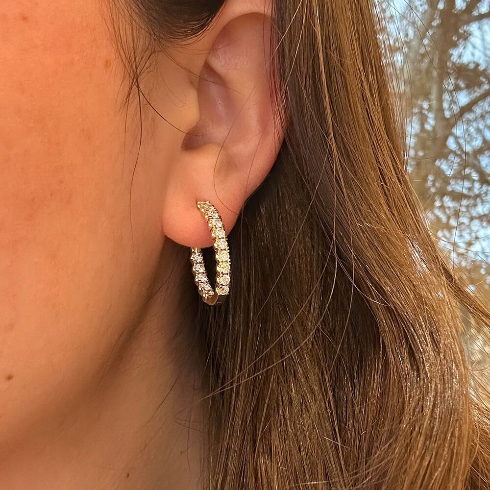 A close-up of a woman's ear wearing a 14K gold hoop earring set with clear diamonds, with brown hair and an outdoor background visible.