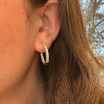 A close-up of a woman's ear wearing a 14K gold hoop earring set with clear diamonds, with brown hair and an outdoor background visible.