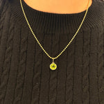 A close-up of a woman wearing a delicate 14K gold chain necklace with a round yellow-green peridot gemstone pendant, styled over a black cable-knit sweater.