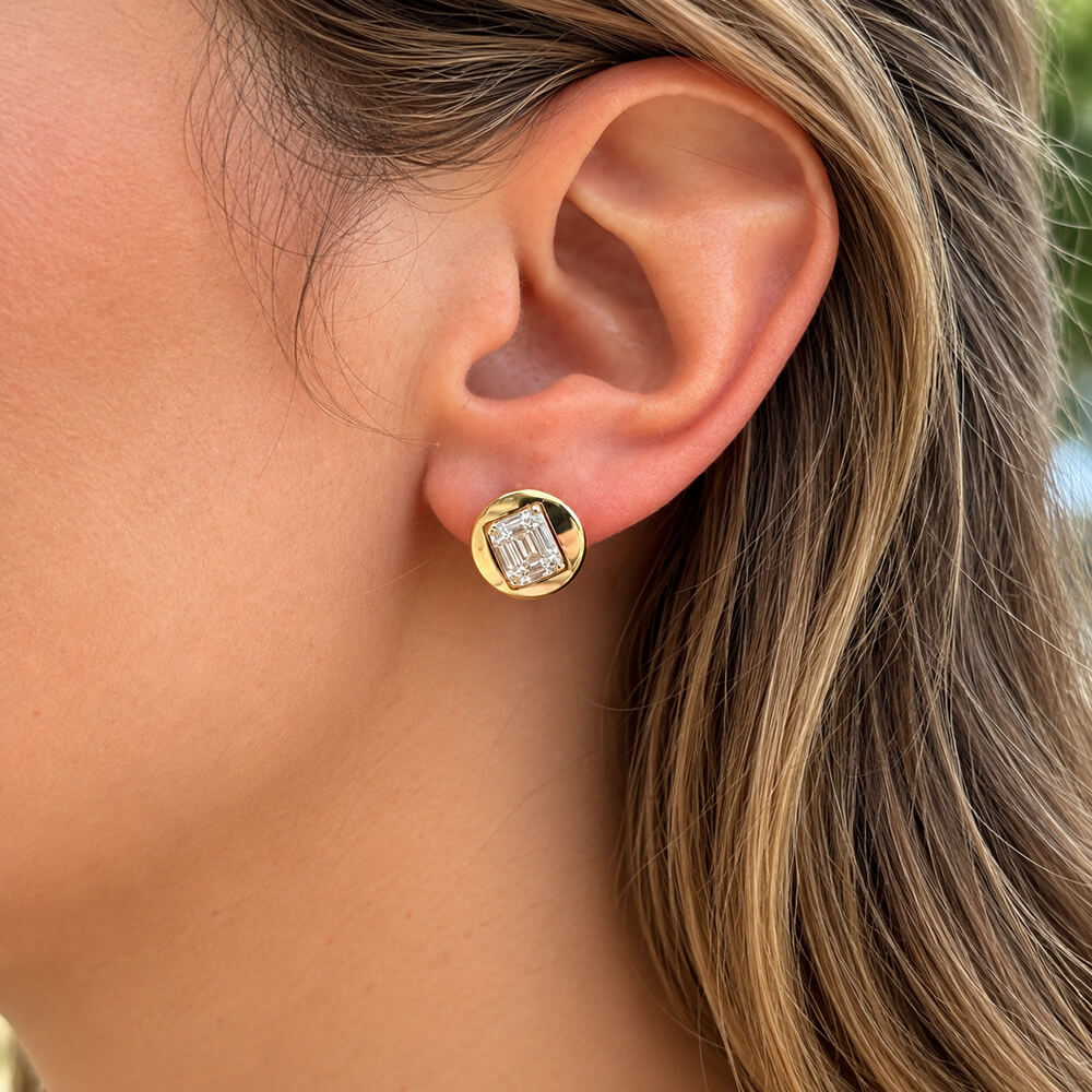 A close-up of a woman's ear wearing a 14k yellow gold stud earring with a clear baguette-cut diamond.