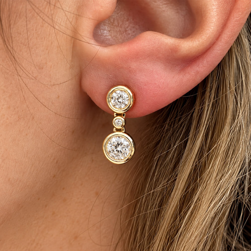A close-up of a woman's ear wearing a gold drop earring with two round clear diamonds, set against skin and blonde hair.