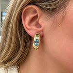 A close-up of a woman's ear wearing a gold hoop earring with a central oval light blue topaz stone, accented by small green periodt stones and clear diamonds.