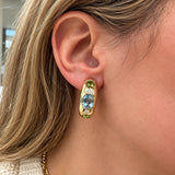 A close-up of a woman's ear wearing a gold hoop earring with a central oval light blue topaz stone, accented by small green periodt stones and clear diamonds.