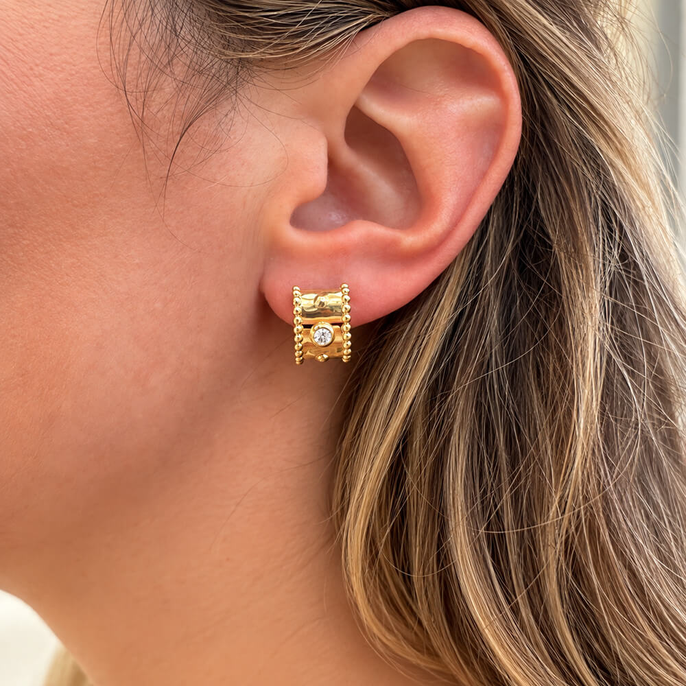 A close-up of a woman's ear wearing a 14k yellow gold huggie hoop earring with a central clear diamond.