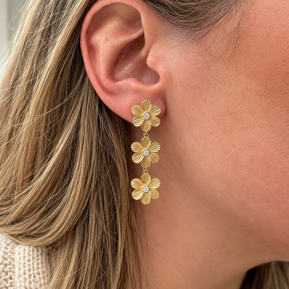 A close-up of a woman's ear wearing a gold dangle earring with three textured flower charms, each set with a small clear diamond.