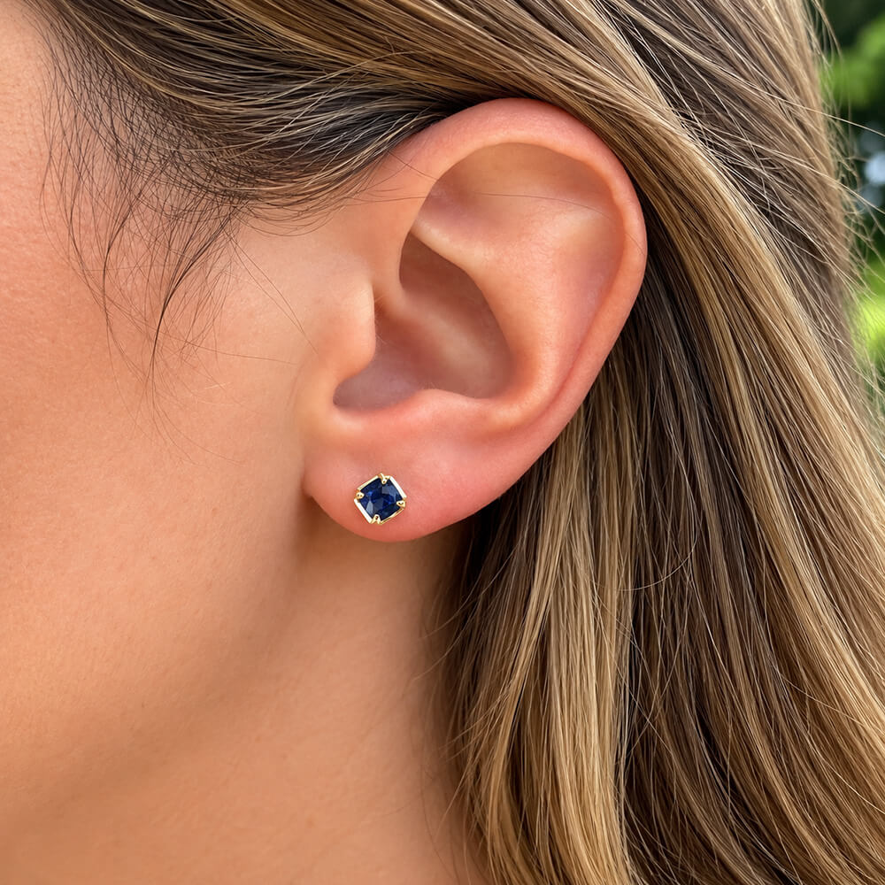 A close-up of a woman's ear wearing a stud earring with a dark blue faceted sapphire in a 14k yellow gold setting.