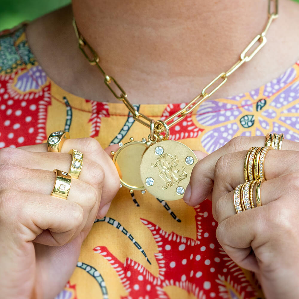 A close-up lifestyle shot of a woman wearing a 14K yellow gold chain necklace with a round pendant and matching gold rings on their fingers, set against a colorful patterned shirt.