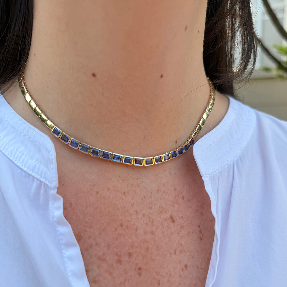 A close-up of a 14K yellow gold tennis-style choker necklace with rectangular blue sapphires, worn by a person wearing a white collared shirt.
