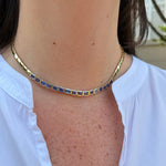 A close-up of a 14K yellow gold tennis-style choker necklace with rectangular blue sapphires, worn by a person wearing a white collared shirt.